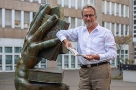 Referent Michael Serrer hält ein Buch in der Hand, angelehnt an eine große Hand-Statue, ebenfalls mit Buch.