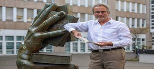 Referent Michael Serrer hält ein Buch in der Hand, angelehnt an eine große Hand-Statue, ebenfalls mit Buch.