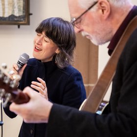 Ein Mann mit Gitarre und eine Frau mit Mikrofon, beide schwarz gekleidet