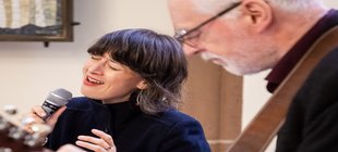 Ein Mann mit Gitarre und eine Frau mit Mikrofon, beide schwarz gekleidet