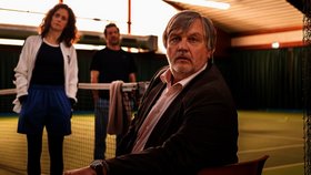 Drei Personen in einer Tennishalle neben einem Tennisnetz; eine Person sitzt im Vordergrund auf einem Stuhl, zwei weitere stehen im Hintergrund auf dem Spielfeld.