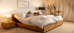 Besser schlafen im Alter Zwei Erwachsene und eine Junge sitzen in einem Bett und schauen ein Buch an