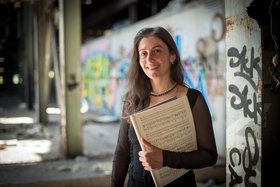 Frau in schwarzer Kleidung mit langen dunklen Haaren und einem Notenheft in der Hand vor einer Wand mit Graffiti