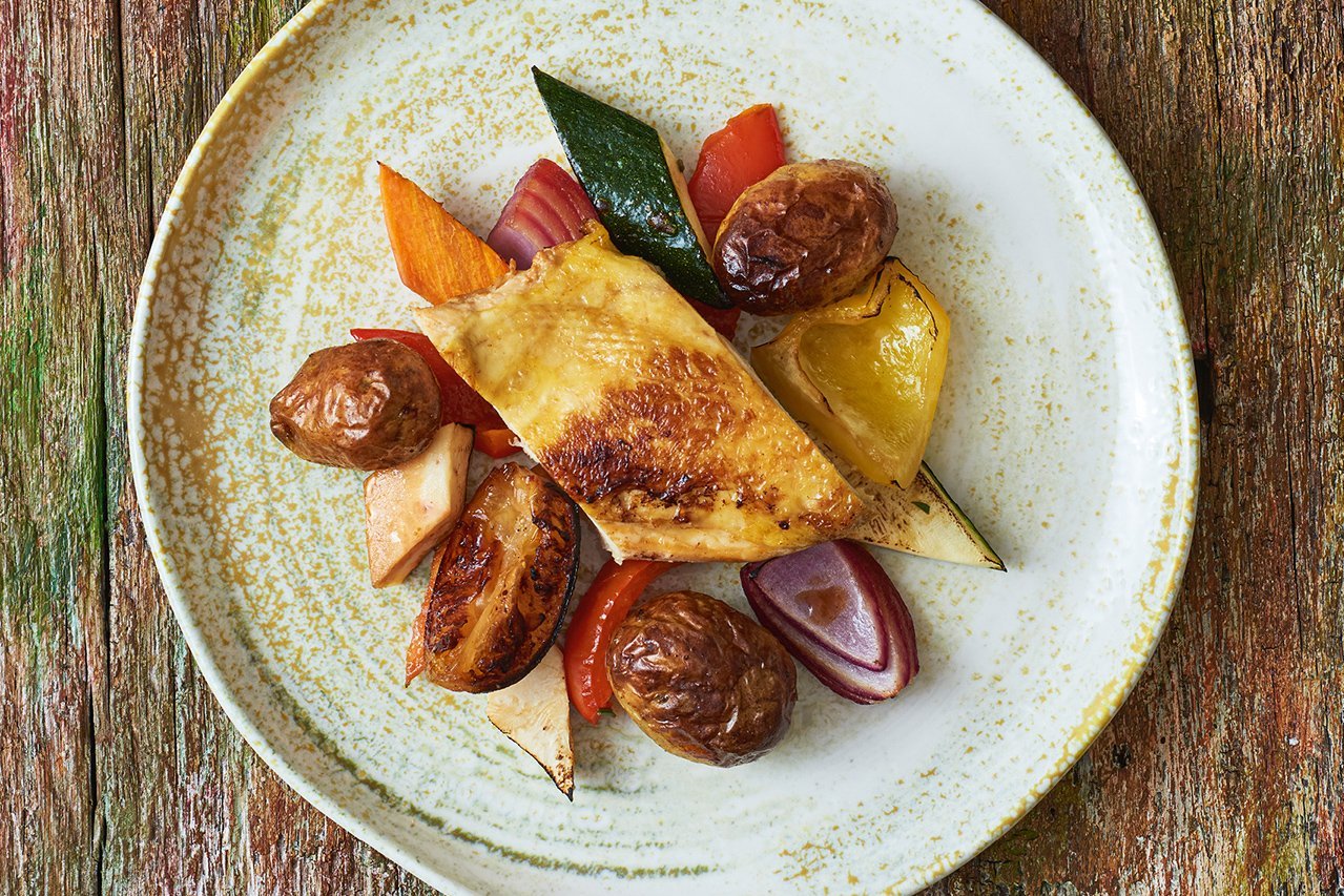 Auf einem weiß-grünen runden Teller, der auf einem grün gespickten Holzuntergrund steht, ist ein Stück Hähnchenbrust mit großen bunten Gemüsestücken (Zucchini, Paprika, Zwiebeln, Kartoffeln) angerichtet.