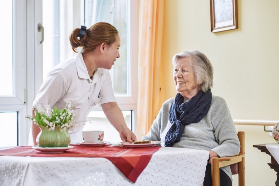Augustinum Berchtesgadener Land Stationaere Pflege Verpflegung Eine Seniorin sitzt an einem Tisch und eine Mitarbeiterin bringt ihr etwas zu essen