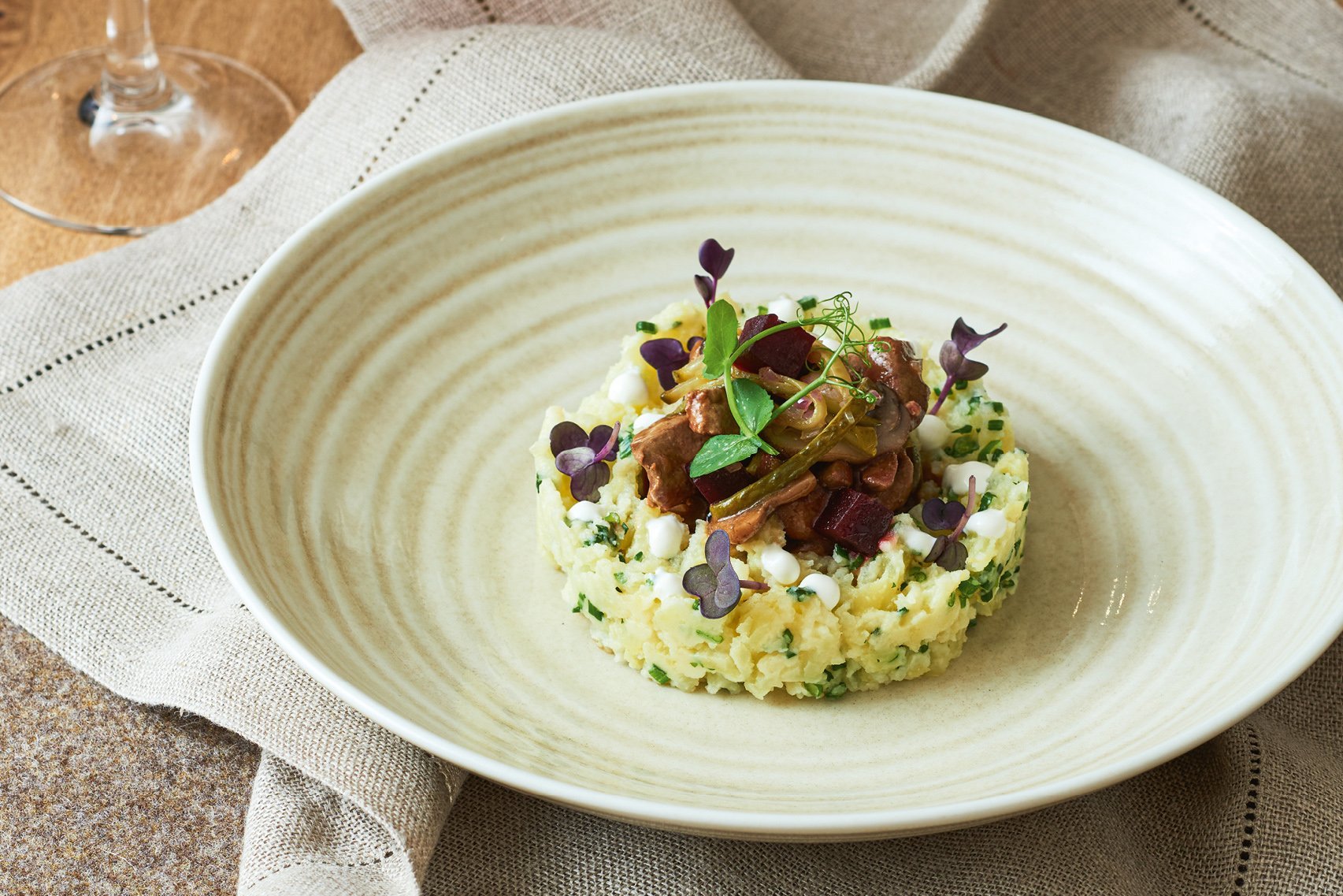 In einem sandfarbenen tiefen Teller ist Kartoffelstampf als Kreis angerichtet, darauf die Rinderleber. Der Teller wird von einer Leinenserviette umrandet, im Hintergrund steht ein Weinglas