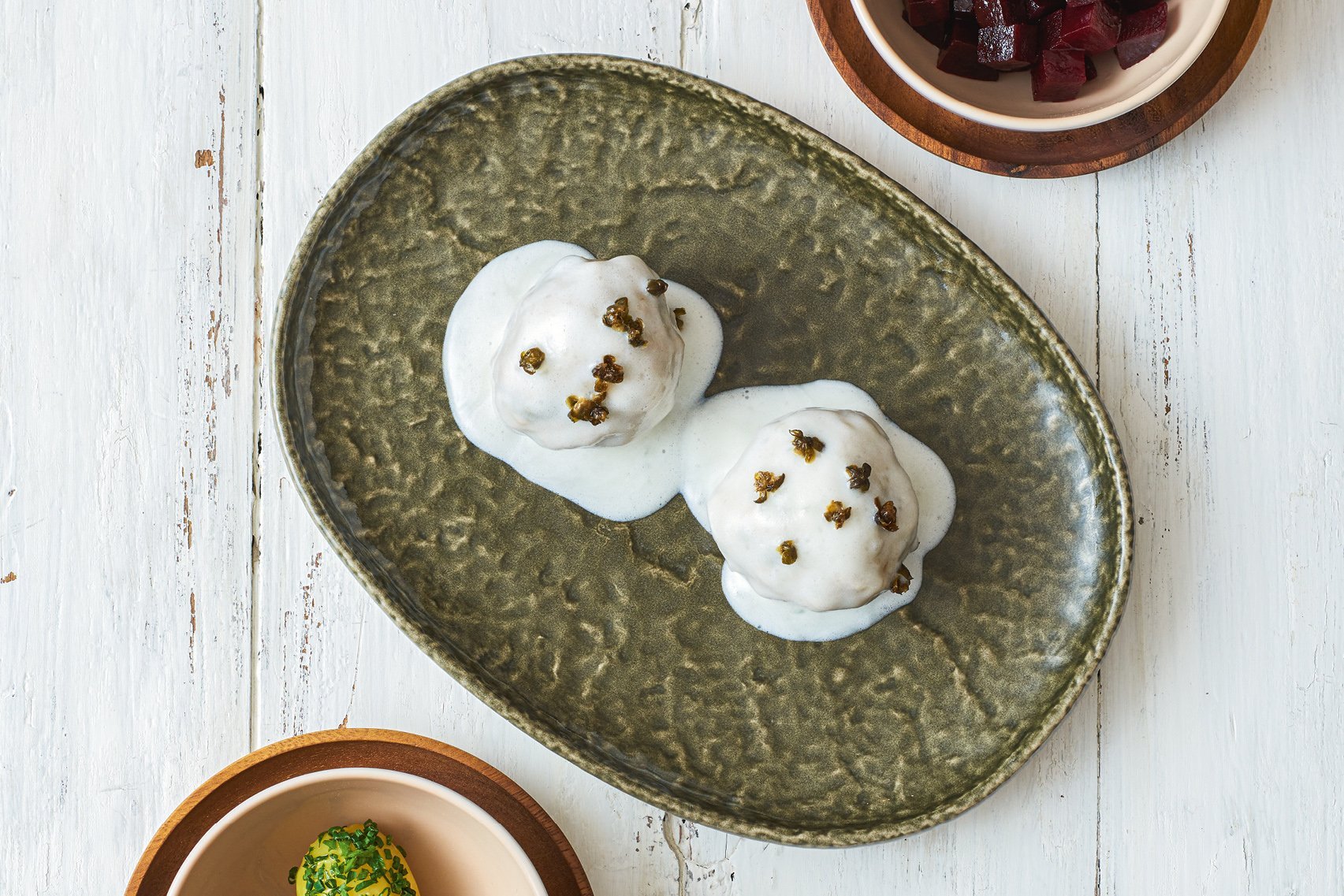 Zwei Königsberger Klopse auf einem grünen Teller angerichtet, als Beilage rote Bete und Kartoffeln auf separaten Tellern 