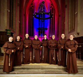 Acht Personen in braunen Ordensgewändern stehen vor dem Altarraum einer Kirche mit farbig beleuchteten Spitzbogenfenstern und Kreuz im Hintergrund
