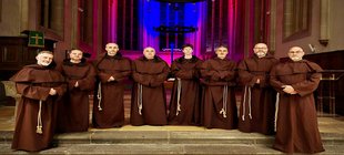 Acht Personen in braunen Ordensgewändern stehen vor dem Altarraum einer Kirche mit farbig beleuchteten Spitzbogenfenstern und Kreuz im Hintergrund
