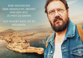 Bärtiger Mann mit Brille blickt geradeaus hinter ihm die Umrisse einer Stadt am Meer