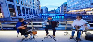 Drei Musiker vor einer Stadtkulisse mit Wasser und Häusern