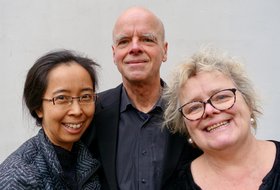 Ein Mann und zwei Frauen in schwarzer Kleidung vor einem grauen Hintergrund