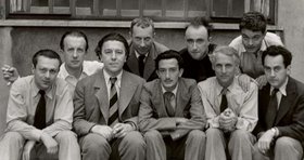 André Breton, Paul Èluard und die Surrealisten André Breton sitz an einem mit Büchern und Papieren beladenen Schreibtisch, mit surrealistischen Werken an der Wand im Hintergrund.
