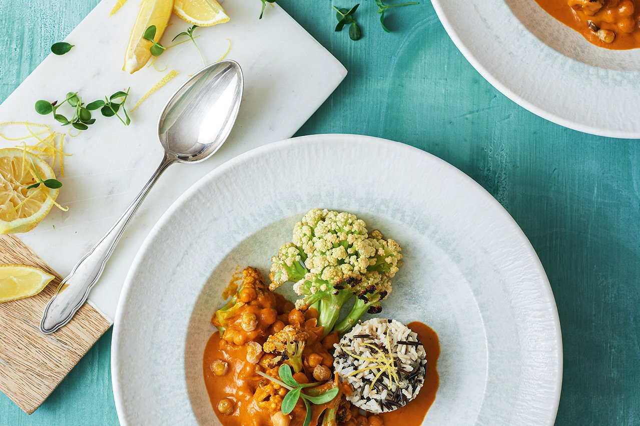 Ein weißer tiefer Teller mit breiter Fahne steht auf einem kräftig türkisen Hintergrund. Es ist wilder Blumenkohl, Reis und eine orangene Soße mit Kichererbsen darauf angerichtet. Links im Bild steht ein weißes Brett mit Zitronenspalten und einemSilberlöffel.