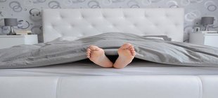 Zwei Füße lugen unter der grauen Bettdecke eines weißen Bettes hervor