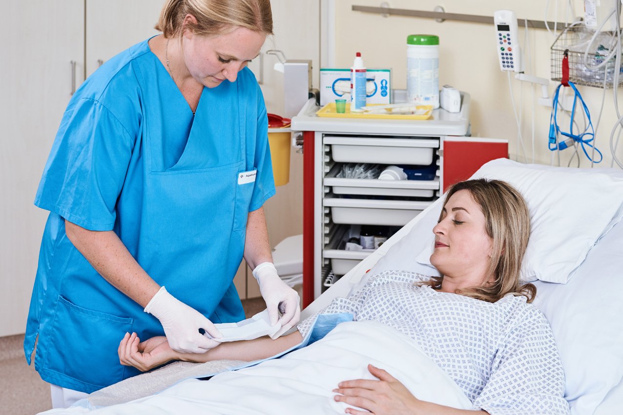 Eine Gesundheits- und Krankenpflegerin legt einer im Bett liegenden Patientin einen Verband am Unterarm an.