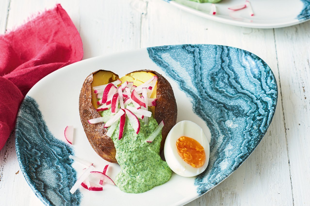 Ein ovaler Teller mit blauen Wellen am Rand steht auf einem weißen Holzhintergrund. Am linken Rand eine pinke Serviette. In der Mitte des Tellers liegt eine aufgeschnittene gebackene Kartoffel gefüllt mir grüner Soße und Radieschenspalten. Dazu ein halbes gekochtes Ei.