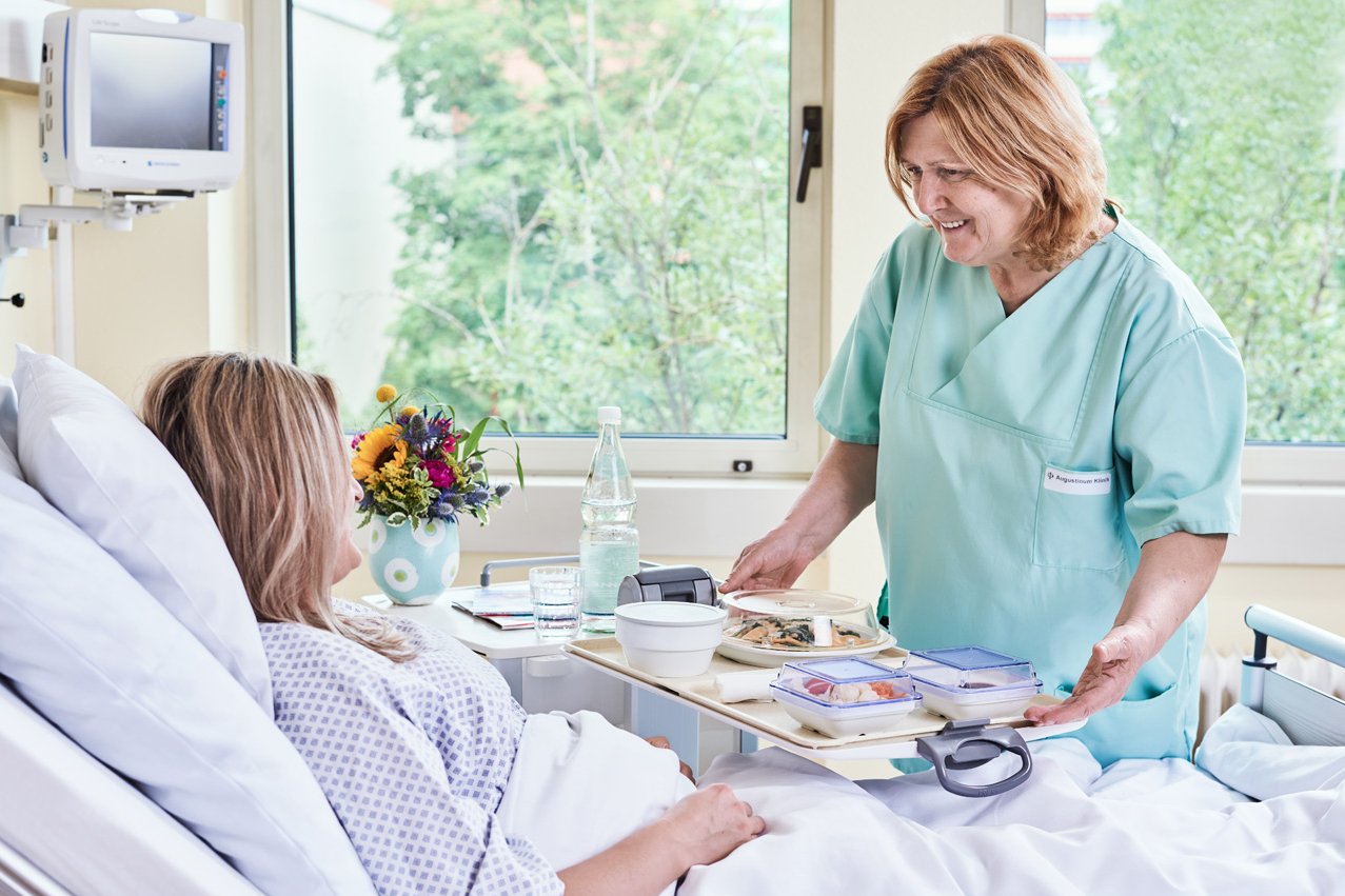 Eine Pflegerin serviert einer im Bett liegenden Patienten auf einem Tablett eine mahlzeit