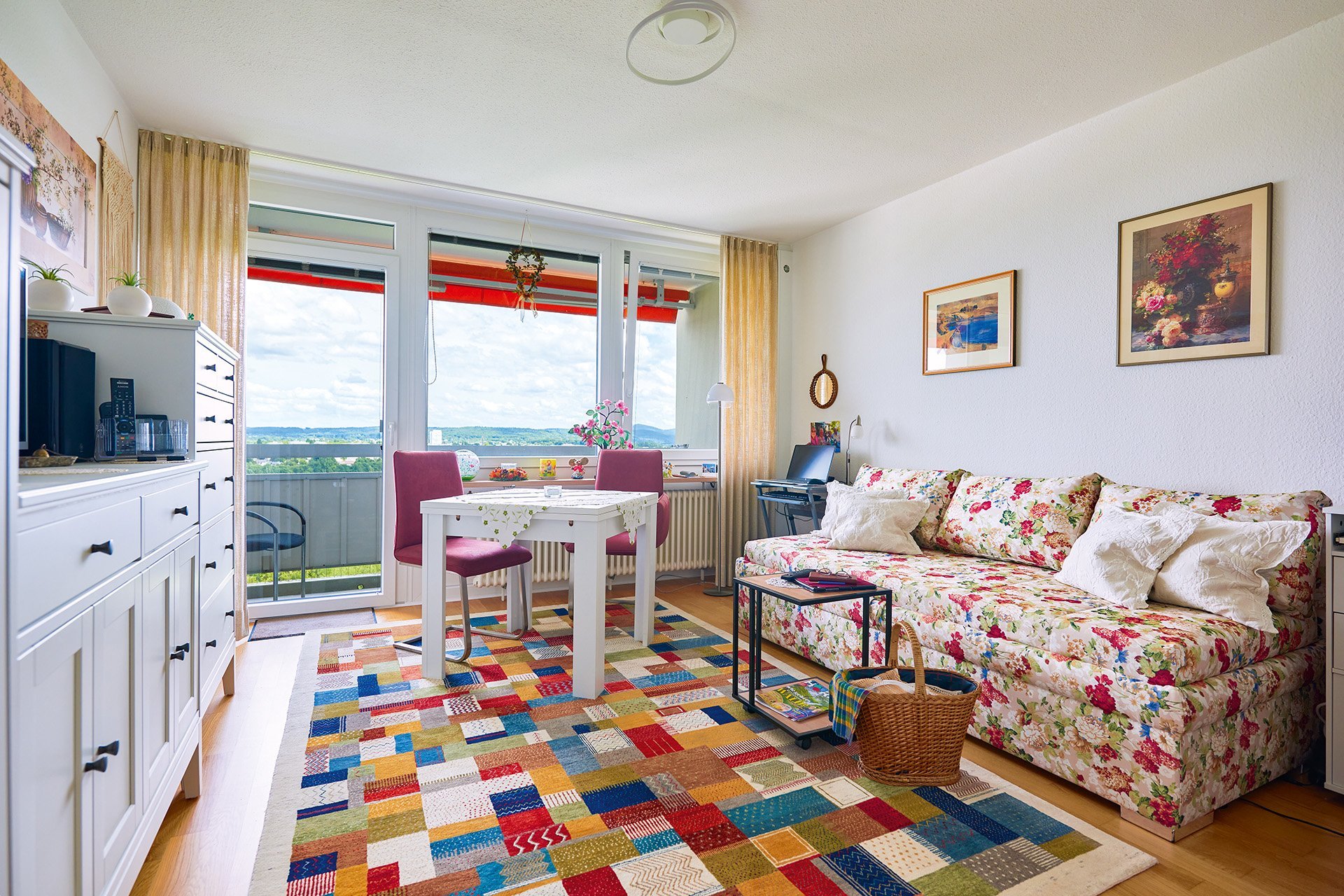 Ein bunt eingerichtetes Wohnzimmer mit einem Sofa, einem kleinen Esstisch, Schränken und einem bunten Teppich