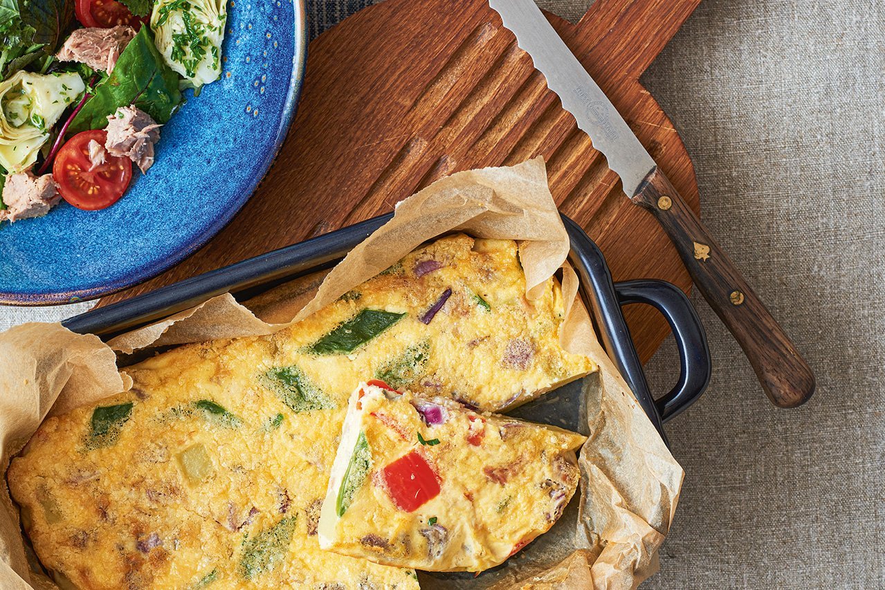 Ein Bräter mit Backpapier und gebackenem Eierkuchen mit Gemüse steht auf einem Holzbrett. Eine Scheibe ist aufgeschnitten. Am linkeren oberen Rand ein Salat mit Tomaten und Thunfisch.