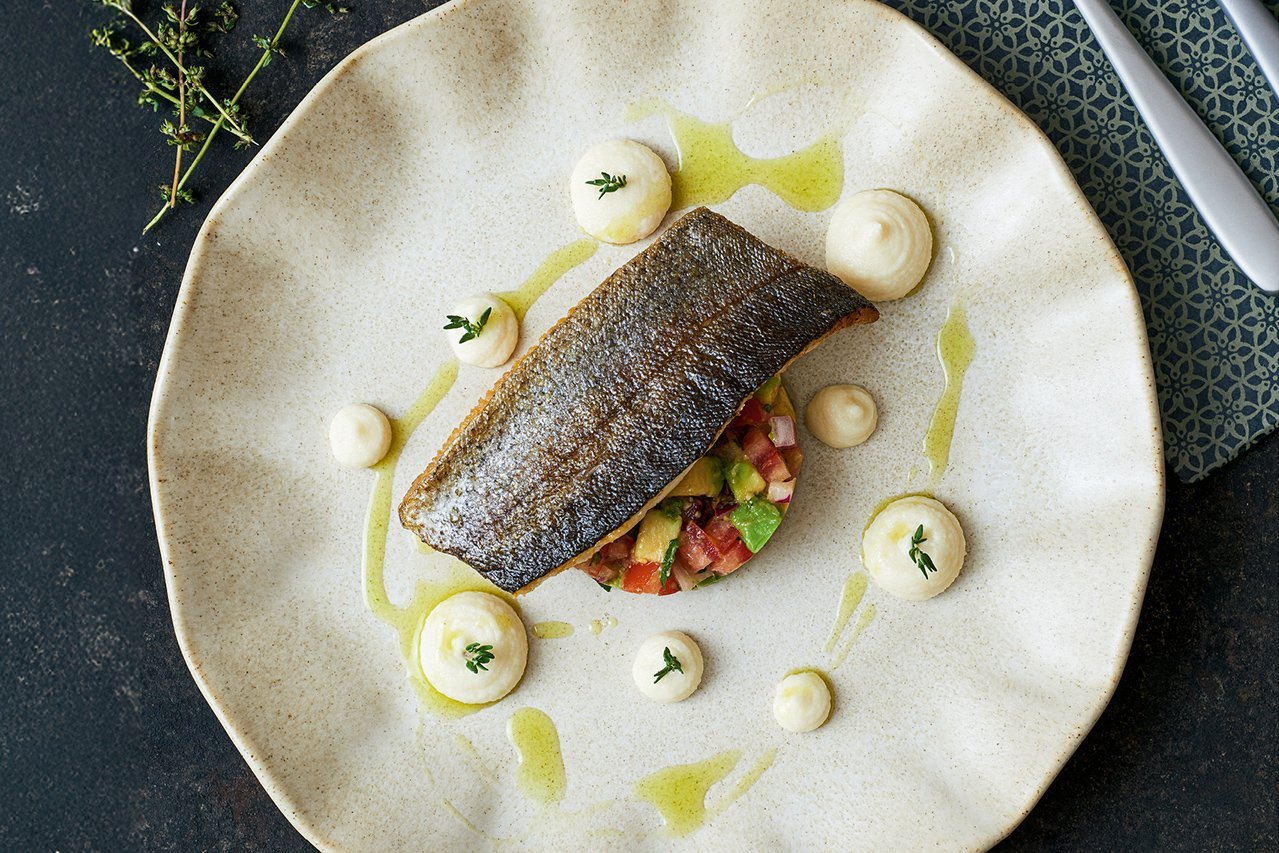 Ein kross gebratenes Forellenfilet liegt auf einem rund ausgestochenen Tomaten-Avocado-Salat, rundherum sind Tupfen von hellem Püree. In der Ecke des Bildes erkennt man frische Thymianzweige