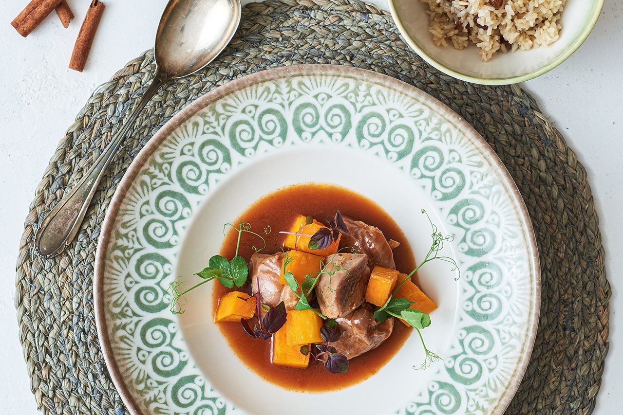 Ein weißer Teller mit grünen Ornamenten auf der breiten Fahne steht auf einem runden Bast-Tischset. Dort angerichtet ist Ragout aus Pute und Kürbis. Man sieht zudem ein
