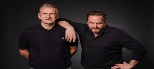 Zwei Männer in schwarzen Hemden vor einem dunklen Hintergrund