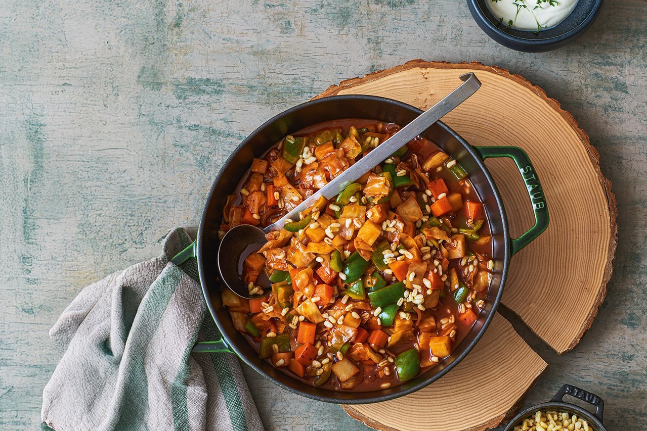 Auf einer rustikalen Holzscheibe steht ein großer Topf mit vegetarischem Gulasch mit großer Kelle. Ein grau-grünes Geschirrtuch liegt bereit um den heißen Topf besser halten zu können.