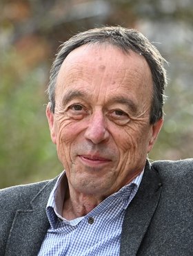 Portraitaufnahme eines älteren Mannes mit dunkeln Haaren vor einem unscharfen Hintergrund. 