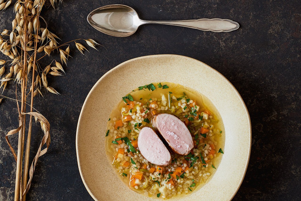 Auf einem schwarzen Blech-Hintergrund steht ein gelblicher runder, tiefer Teller. Darüber liegt quer ein Löffel. Links neben dem Teller liegen zwei Stiele reifer Hafer. Im Teller ist ein Graupen-Gemüse-Sud angerichtet, darin liegen zwei Stücke Schweinefilet.
