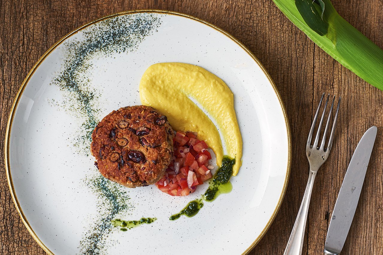 Auf einem edlen weißen Teller mit Goldrand und dunkelgrünen Tupfen, der auf einem Holzuntergrund steht, ist kräftig gelbes Maispüree mit Tomatensalsa und einem Bohnenbratling angerichtet. An der Seite liegt junges Maiskolben-Grün.