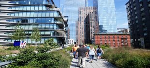 Menschen spazieren auf einem begrünten Fußweg zwischen modernen Hochhäusern und historischen Backsteingebäuden in im dicht bebauten New York.