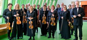 Die Musiker*innen des Kammerorchesters Arcata stehen mit ihren Instrumenten in der Hand in schwarzer Konzertkleidung vor einer Fensterfront.