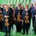 Ein Abend voller Operndramatik und Streicherglanz Die Musiker*innen des Kammerorchesters Arcata stehen mit ihren Instrumenten in der Hand in schwarzer Konzertkleidung vor einer Fensterfront.