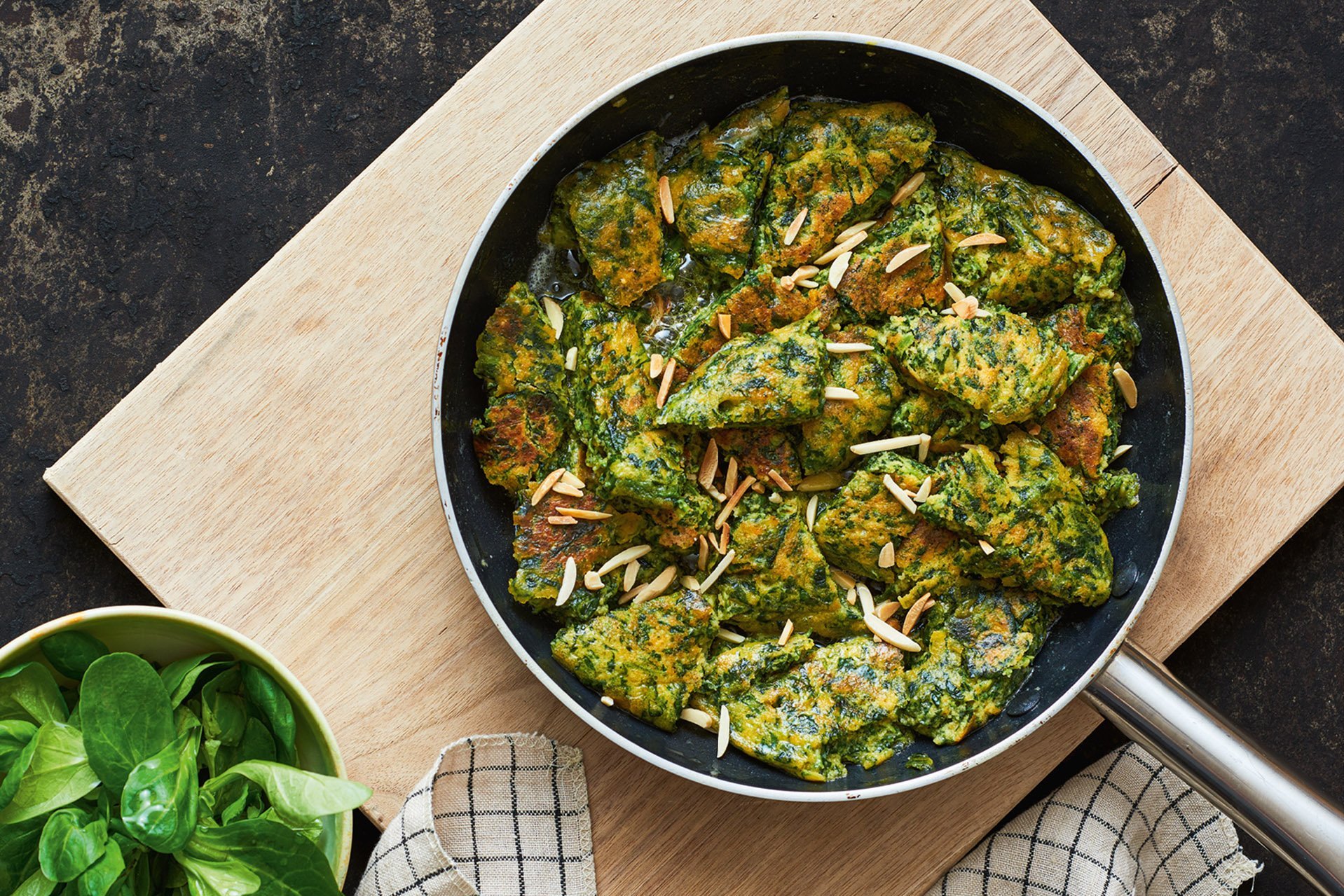 Auf einem Holzbrett steht eine beschichtete Pfanne mit gelb-grünem Schmarren. Am Rand ist ein Beilagensalat zu erkennen.