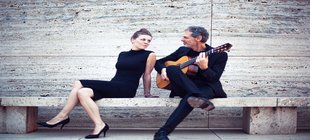 Eine Musikerin und ein Musiker mit Gitarre auf einer Bank vor einer Steinwand