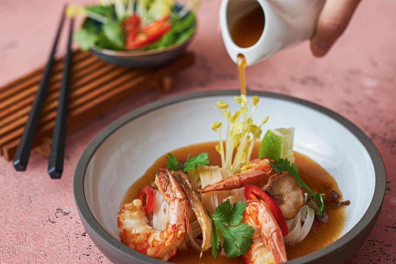 Suppe wird in einen tiefen weißen Teller mit schwarzem Rand eingegossen. Im Teller befinden sich Garnelen, Reisnudeln, Chili, Pilze und Koriander. Im Hintergrund steht ein Brett mit Kräutern und Stäbchen.