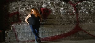 Frau mit langen roten Haaren in Jeans und schwarzem Top vor einer Wand mit Graffiti