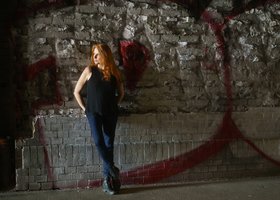 Frau mit langen roten Haaren in Jeans und schwarzem Top vor einer Wand mit Graffiti
