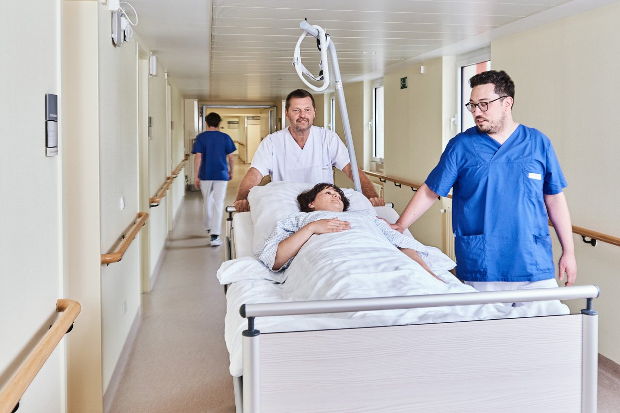 Zwei Pfleger schieben ein Bett über den Gang, in dem eine Patientin liegt.