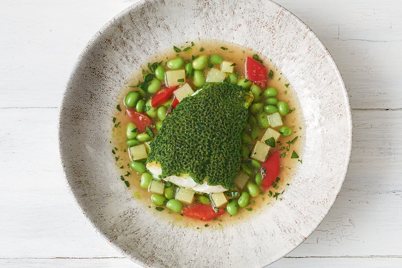 Ein weiß-grauer runder Teller steht auf weißem Holz, keine weitere Dekoration im Bild. In der Mitte ein Bohnen-Sud mit roter Paprika, darauf ein Stück fisch mit grasgrüner Kräuterkruste.