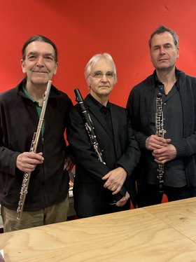 Drei schwarzgekleidete Männer mit Flöte, Oboe und Klarinette vor rotem Hintergrund