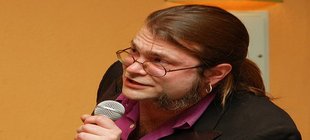 Ein Mann mit Brille und Mikrofon vor einem ockerfarbenen Hintergrund
