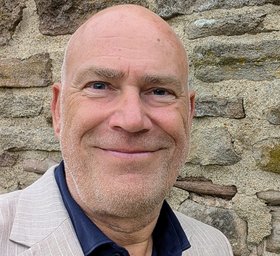 Ein Mann in dunklem Hemd und hellem Anzug vor einer Steinmauer