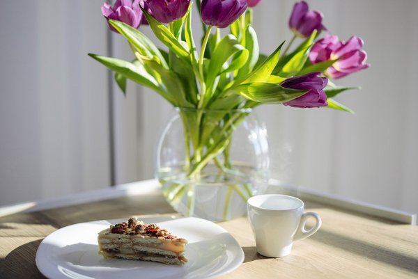 Auf einem Tisch steht ein Strauß violetter Tulpen neben einem Stück Kuchen und einer Tasse Kaffee