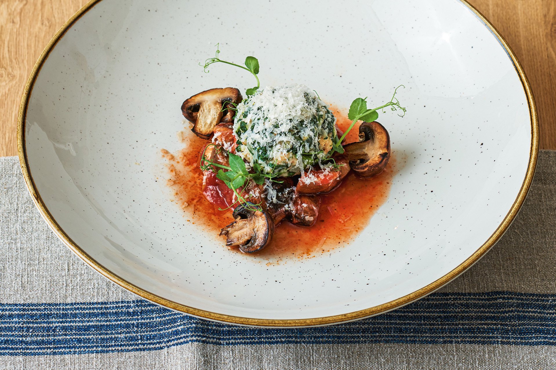 In einem großen tiefen weißen Teller liegt ein Spinatknödel mit viel Käse, dekoriert mit mit Pilzen und Tomatenragout. 