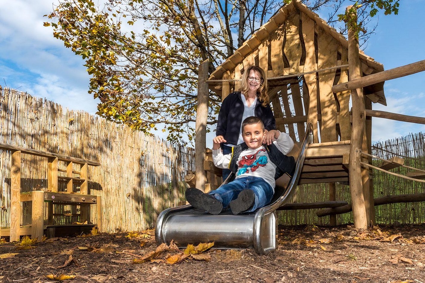 fröhlicher Junge auf einer Rutsche, im Hintergrund eine Erzieherin