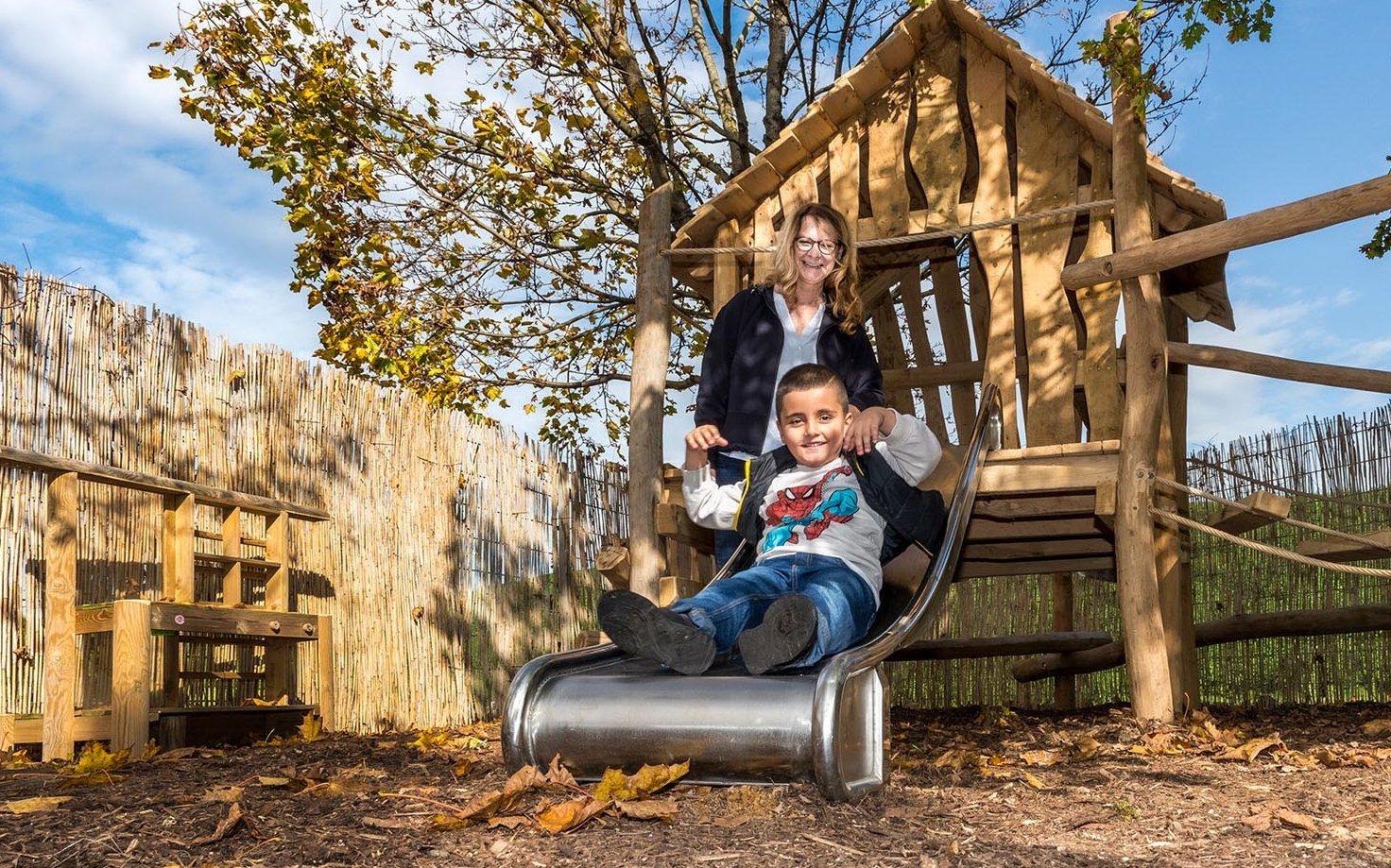 fröhlicher Junge auf einer Rutsche, im Hintergrund eine Erzieherin