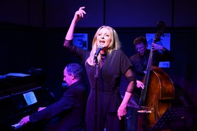 Sängerin an einem Mikrofon während eines Jazzkonzerts, begleitet von einem Pianisten am Flügel und einem Kontrabassisten auf einer stimmungsvoll beleuchteten Bühne.