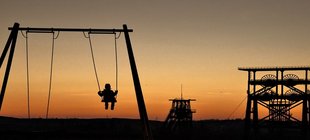 Kind auf einer Schaukel im Abendlicht mit Hochofen im Hintergrund